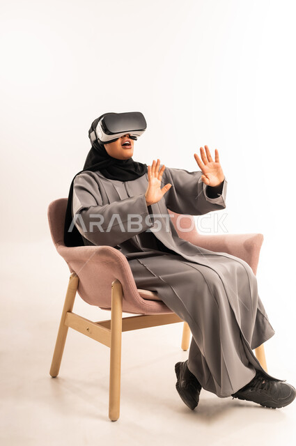 التفاعل والاندماج مع الواقع الافتراضي، محاولة ابعاد وإيقاف شيء ما، محاكاة العالم الخيالي، بورتريه لشابة عربية خليجية سعودية محجبة تجلس على كرسي مريح ترتدي العباءة ونظارة في آر ثلاثية الأبعاد ترفع يديها بحماس بتعابير المفاجأة، خلفية بيضاء
