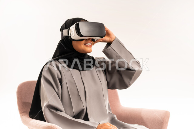 الاستمتاع بمشاهدة أفلام الواقع الافتراضي ومحاكاة العالم الخيالي, التطور التقني والتكنولوجي, قضاء وقت الفراغ بالنشاط الترفيهي, بورتريه مقرب لامرأة عربية خليجية سعودية محجبة تجلس على كرسي مريح ترتدي العباءة و نظارة في آر ثلاثية الأبعاد بتعابير السرور, خلفية بيضاء