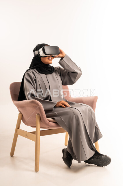 Technical and technological development, enjoying watching virtual reality movies and simulating the imaginary world, spending free time with recreational activities, portrait of a veiled Saudi Arabian Gulf woman sitting on a comfortable chair wearing an abaya and 3D VR glasses with expressions of pleasure, white background