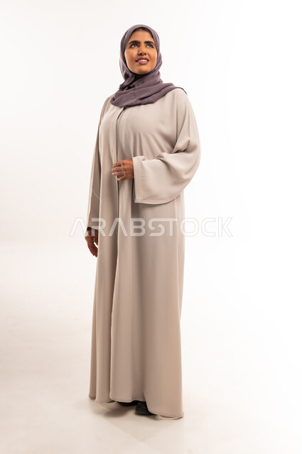 Concern for elegance and appearance, concept of femininity and softness, portrait of a smiling veiled Saudi Arabian Gulf woman wearing a gray abaya standing straight and looking at something above with gestures of happiness and pleasure, full length body, white background