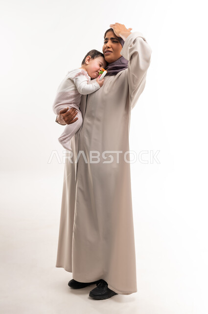 The role of mother in raising children, spending time with infants, the concept of motherhood and caring for and nurturing children, feeling helpless and ignorant, portrait of a veiled Saudi Arabian Gulf woman wearing a gray abaya holding her infant and putting her hand on her head, full-length body, white background