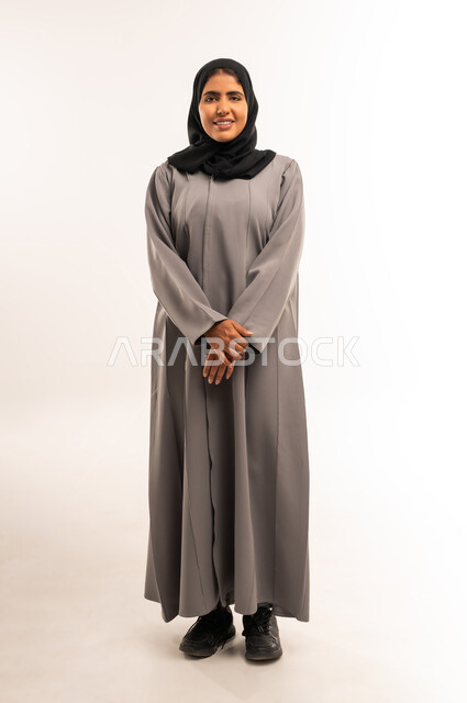 Concern for elegance and appearance, concept of femininity and softness, portrait of a smiling veiled Saudi Arabian Gulf woman wearing a gray abaya standing straight and looking at the camera with gestures of happiness and pleasure, full body, white background