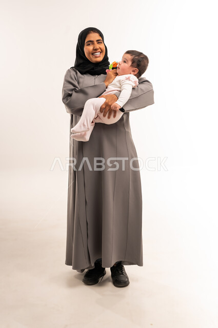 حنان الأمهات و رعايتهم للأولاد, مداعبة الأم لطفلتها بإيماءات السعادة, الاستمتاع بقضاء الوقت الممتع مع الأبناء, بورتريه لامرأة عربية خليجية سعودية محجبة ترتدي عباءة رمادية تقوم باللعب مع رضيعتها, الجسم بالطول الكامل, خلفية بيضاء