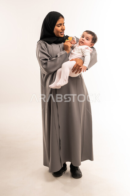 مداعبة الأم لطفلتها بإيماءات السعادة, حنان الأمهات و رعايتهم للأولاد, الاستمتاع بقضاء الوقت الممتع مع الأبناء, بورتريه لامرأة عربية خليجية سعودية محجبة ترتدي عباءة رمادية تقوم باللعب مع رضيعتها, الجسم بالطول الكامل, خلفية بيضاء
