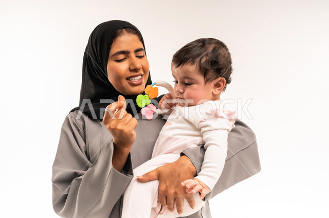 الاستمتاع بقضاء الوقت الممتع مع الأبناء, مداعبة الأم لطفلتها بإيماءات السعادة, حنان الأمهات و رعايتهم للأولاد, بورتريه مقرب لامرأة عربية خليجية سعودية محجبة ترتدي عباءة رمادية تقوم باللعب مع رضيعتها, خلفية بيضاء