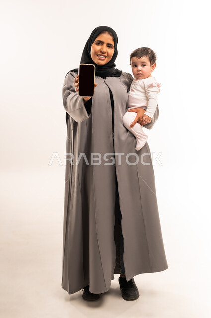 مفهوم الأمومة والعناية بالطفل, استخدام جهاز تقني حديث ومتطور, النظر إلى الكاميرا بإيماءات السعادة والسرور, بورتريه لامرأة عربية خليجية سعودية محجبة ترتدي عباءة رمادية تحمل رضيعتها وتمسك بيدها الهاتف المحمول, عرض شاشة سوداء فارغة عبر الجوال, خلفية بيضاء