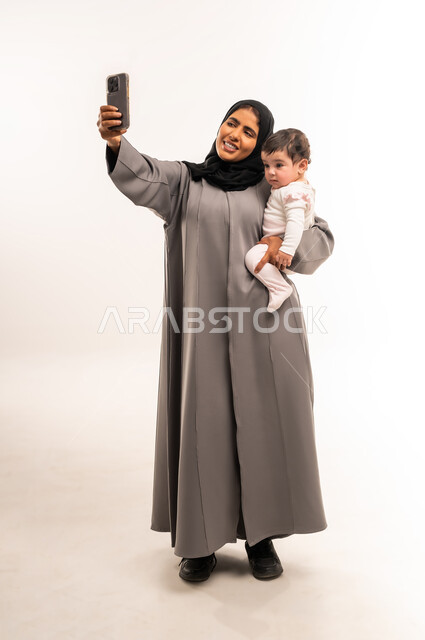 Documenting happy moments with children, using a modern and advanced technical device, the concept of motherhood and child care, portrait of a veiled Saudi Arabian Gulf woman wearing a gray abaya holding her baby and taking a selfie with a mobile phone, full body, white background