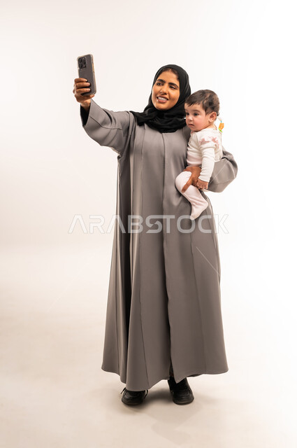 Using a modern and advanced technical device, documenting happy moments with children, the concept of motherhood and child care, portrait of a veiled Saudi Arabian Gulf woman wearing a gray abaya holding her baby and taking a selfie with a mobile phone, full body, white background