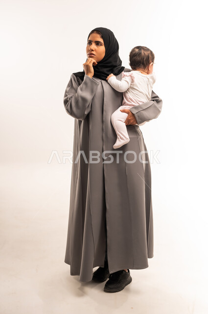 التأمل والتخطيط للمستقبل, مفهوم الأمومة والطفولة, تعابير شرود الذهن والتفكير, دور الأم في رعاية الأبناء, بورتريه لامرأة عربية خليجية سعودية محجبة ترتدي عباءة رمادية تحمل رضيعتها وتضع يدها على ذقنها بإيماءات التركيز والتمعن, الجسم بالطول الكامل, خلفية بيضاء