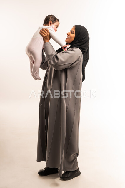 Mother caressing her child with gestures of happiness, enjoying spending quality time with children, mothers' tenderness and care for children, portrait of a veiled Saudi Arabian Gulf woman wearing a gray abaya playing with her baby, full body, white background