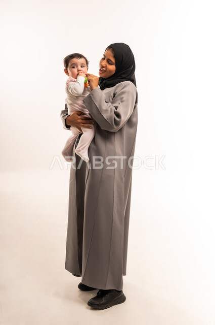 الاستمتاع بقضاء الوقت الممتع مع الأبناء, مداعبة الأم لطفلتها بإيماءات السعادة, حنان الأمهات و رعايتهم للأولاد, بورتريه لامرأة عربية خليجية سعودية محجبة ترتدي عباءة رمادية تقوم باللعب مع رضيعتها, الجسم بالطول الكامل, خلفية بيضاء