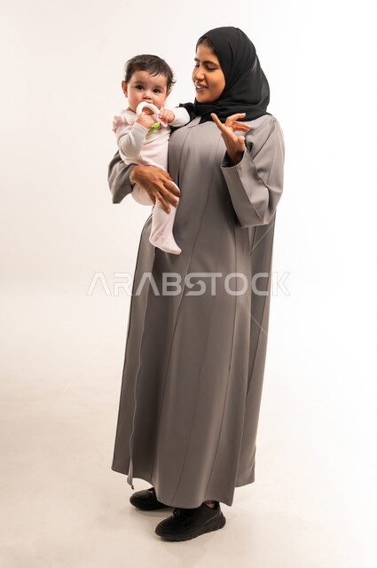 رفع اليد بتعابير التساؤل والاستغراب, الاهتمام والعناية بالطفل, دور الأم في رعاية الأبناء, يورتريه لامرأة عربية خليجية سعودية محجبة ترتدي عباءة رمادية تحمل رضيعتها وترفع يدها بتعابير الاستفهام, الجسم بالطول الكامل, خلفية بيضاء