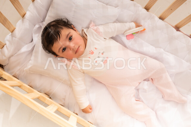 الاهتمام والاعتناء بمظهر الأطفال الخارجي, الملامح الطفولية البريئة الناعمة, مفهوم البراءة والطفولة, صورة علوية عامودية لرضيعة عربية خليجية سعودية ترتدي لباس قطني مريح تستلقي على ظهرها في السرير