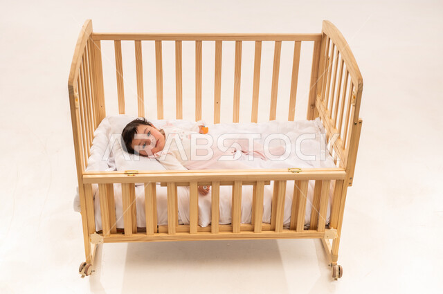 الملامح الطفولية البريئة الناعمة, الاهتمام والاعتناء بمظهر الأطفال الخارجي, مفهوم البراءة والطفولة, بورتريه لرضيعة عربية خليجية سعودية ترتدي لباس قطني مريح تستلقي على ظهرها في السرير, خلفية بيضاء