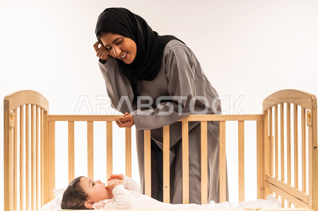 تحمل مسؤولية الاعتناء والاهتمام بالطفل, مفهوم الأمومة والطفولة, الاستمتاع بقضاء وقت ممتع مع الأبناء, بورتريه لامرأة عربية خليجية سعودية محجبة ترتدي عباءة رمادية تتكئ على سرير رضيعتها وتنظر إليها بإيماءات الامتنان والسرور, خلفية بيضاء