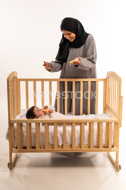 حنان الأمهات و رعايتهم للأولاد, مداعبة الأم لطفلتها بإيماءات السعادة, الاستمتاع بقضاء الوقت الممتع مع الأبناء, بورتريه لامرأة عربية خليجية سعودية محجبة ترتدي عباءة رمادية تقوم باللعب مع رضيعتها, الجسم بالطول الكامل, خلفية بيضاء