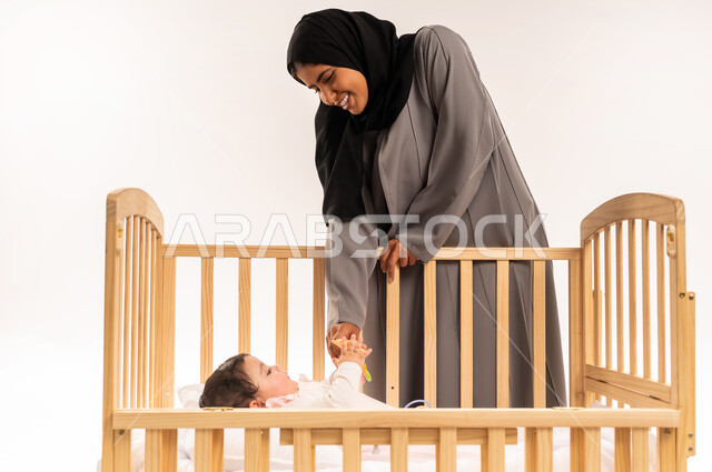 الاستمتاع بقضاء الوقت الممتع مع الأبناء, مداعبة الأم لطفلتها بإيماءات السعادة, حنان الأمهات و رعايتهم للأولاد, بورتريه لامرأة عربية خليجية سعودية محجبة ترتدي عباءة رمادية تقوم باللعب مع رضيعتها, خلفية بيضاء