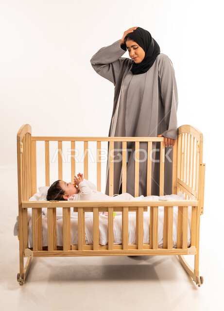 الشعور بالعجز وعدم المعرفة, مفهوم الأمومة والاعتناء والرعاية بالطفل, دور الأم في تربية الأبناء, قضاء الوقت مع الرضع, بورتريه لامرأة عربية خليجية سعودية محجبة ترتدي عباءة رمادية تقف أمام سرير رضيعتها و تضع يدها على رأسها, الجسم بالطول الكامل, خلفية بيضاء
