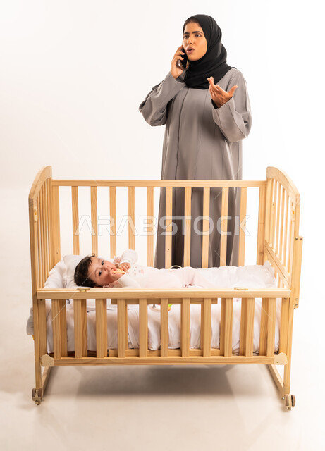 رفع اليد بتعابير التساؤل والاستفهام, استخدام جهاز تقني حديث ومتطور, الاهتمام والعناية بالطفل, بورتريه لامرأة عربية خليجية سعودية محجبة ترتدي عباءة رمادية تقف أمام سرير الرضيعة وتقوم بإجراء مكالمة هاتفية عبر الجوال, الجسم بالطول الكامل, خلفية بيضاء
