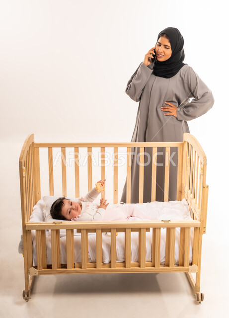 إجراء مكالمة هاتفية عبر الجوال, مفهوم الأمومة والطفولة, استخدام جهاز تقني حديث ومتطور, الاهتمام والعناية بالطفل, بورتريه لامرأة عربية خليجية سعودية محجبة ترتدي عباءة رمادية تقف أمام سرير الرضيعة وتضع يدها على خصرها, الجسم بالطول الكامل, خلفية بيضاء