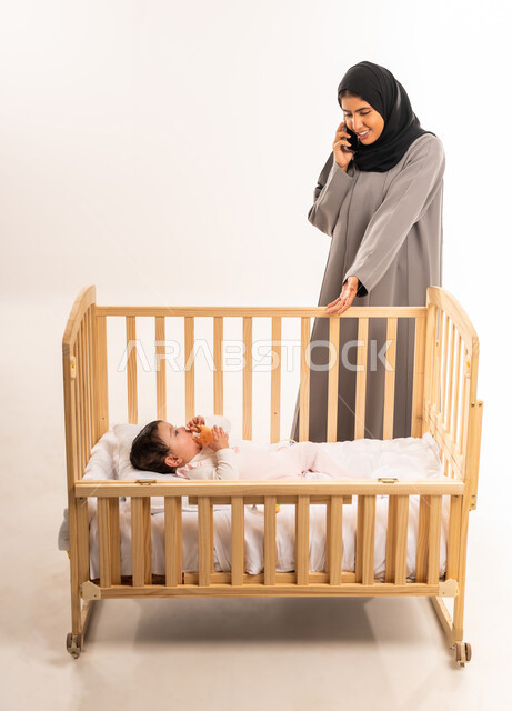 الاهتمام والعناية بالطفل, مفهوم الأمومة والطفولة, استخدام جهاز تقني حديث ومتطور, بورتريه لامرأة عربية خليجية سعودية محجبة ترتدي عباءة رمادية تشير بيدها إلى رضيعتها وتقوم بإجراء مكالمة هاتفية عبر الجوال, الجسم بالطول الكامل, خلفية بيضاء