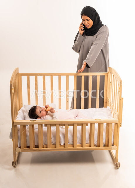 Motherhood and childhood concept, using a modern and advanced technical device, caring for and taking care of the child, portrait of a veiled Saudi Arabian Gulf woman wearing a gray abaya pointing to her infant and making a phone call via mobile, full body, white background