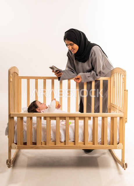 استخدام جهاز تقني حديث ومتطور, توثيق اللحظات السعيدة مع الأبناء, الاستمتاع بقضاء وقت ممتع مع الطفل, بورتريه لامرأة عربية خليجية سعودية محجبة ترتدي عباءة رمادية تقوم بالتقاط صورة لرضيعتها عبر الجوال, الجسم بالطول الكامل, خلفية بيضاء