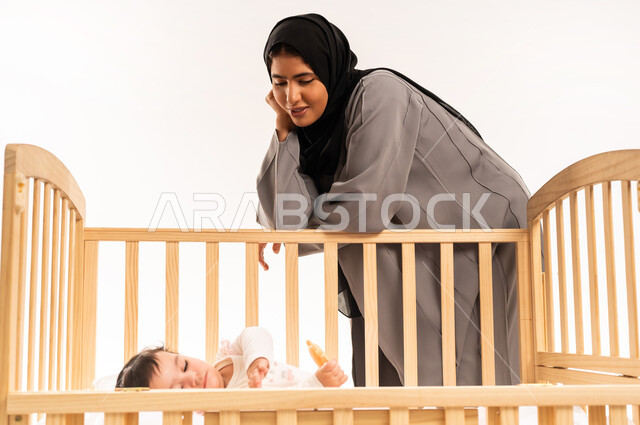 تحمل مسؤولية الاعتناء والاهتمام بالطفل, مفهوم الأمومة والطفولة, الاستمتاع بقضاء وقت ممتع مع الأبناء, بورتريه لامرأة عربية خليجية سعودية محجبة ترتدي عباءة رمادية تتكئ على سرير رضيعتها وتنظر إليها بإيماءات الامتنان والسرور, خلفية بيضاء