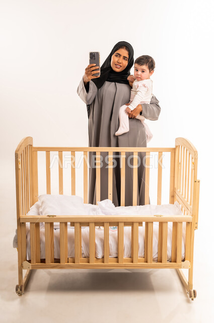 استخدام جهاز تقني حديث ومتطور, توثيق اللحظات السعيدة مع الأبناء, مفهوم الأمومة والعناية بالطفل, بورتريه لامرأة عربية خليجية سعودية محجبة ترتدي عباءة رمادية تحمل رضيعتها وتقوم بالتقاط صورة سيلفي عبر الجوال, الجسم بالطول الكامل, خلفية بيضاء