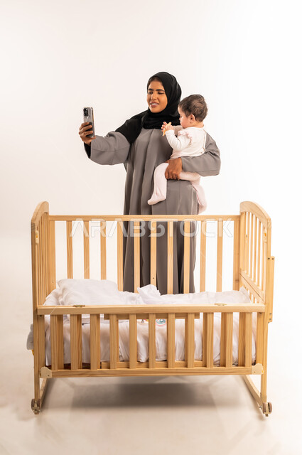 Documenting happy moments with children, the concept of motherhood and child care, using a modern and advanced technical device, portrait of a veiled Saudi Arabian Gulf woman wearing a gray abaya holding her baby and taking a selfie with a mobile phone, full body, white background