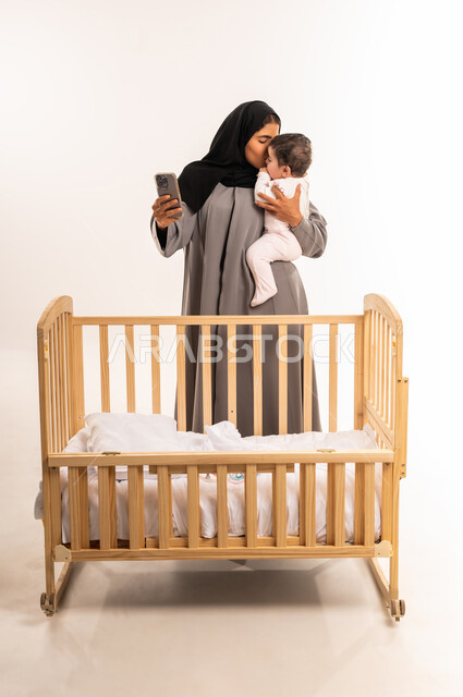 The concept of motherhood and child care, documenting happy moments with children, using a modern and advanced technical device, portrait of a veiled Saudi Arabian Gulf woman wearing a gray abaya taking a selfie with her mobile phone while kissing her baby, full body, white background