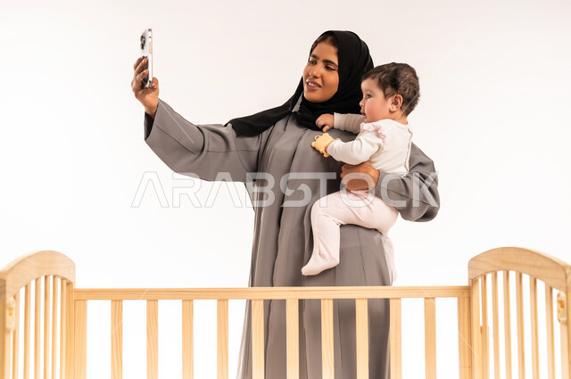 Using a modern and advanced technical device, documenting happy moments with children, the concept of motherhood and child care, a portrait of a veiled Saudi Arabian Gulf woman wearing a gray abaya, holding her baby and taking a selfie with her mobile phone, white background