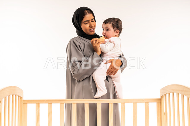 تعزيز رابط الأمومة بين الأم وصغيرتها, الشعور بالأمان والاطمئنان، مفهوم حنان الأمهات و تحمل المسؤولية والعناية بالطفل, بورتريه لامرأة عربية خليجية سعودية محجبة ترتدي عباءة رمادية تقف أمام السرير وتحمل رضيعتها وتنظر إليها بتعابير الحب, خلفية بيضاء