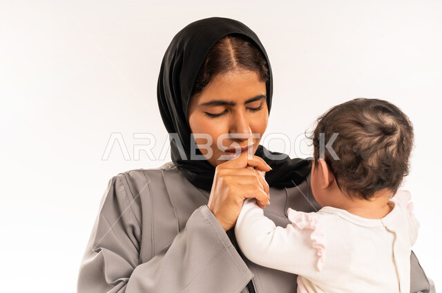 Feeling safe and secure, the concept of maternal tenderness, taking responsibility and caring for children, strengthening the bond of motherhood between mother and child, close-up portrait of a veiled Saudi Arabian Gulf woman wearing a gray abaya holding her infant and kissing his hand with expressions of love, white background