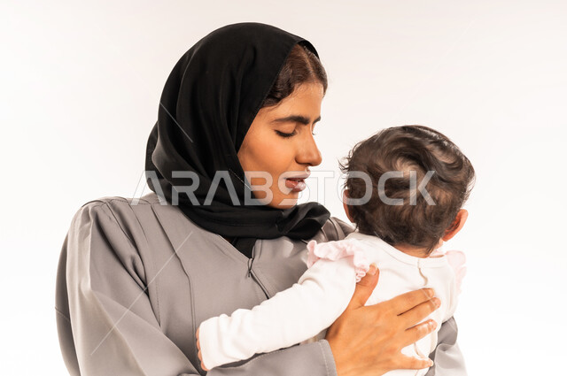 مفهوم حنان الأمهات و تحمل المسؤولية والعناية بالطفل, الشعور بالأمان والاطمئنان, تعزيز رابط الأمومة بين الأم وصغيرتها, بورتريه مقرب لامرأة عربية خليجية سعودية محجبة ترتدي عباءة رمادية تحمل رضيعتها وتنظر إليها بتعابير الحب, خلفية بيضاء
