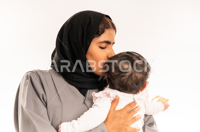 تعزيز رابط الأمومة بين الأم وصغيرتها, الشعور بالأمان والاطمئنان، مفهوم حنان الأمهات و تحمل المسؤولية والعناية بالطفل, بورتريه مقرب لامرأة عربية خليجية سعودية محجبة ترتدي عباءة رمادية تحمل رضيعتها وتقوم بتقبيل رأسها بتعابير الحب, خلفية بيضاء