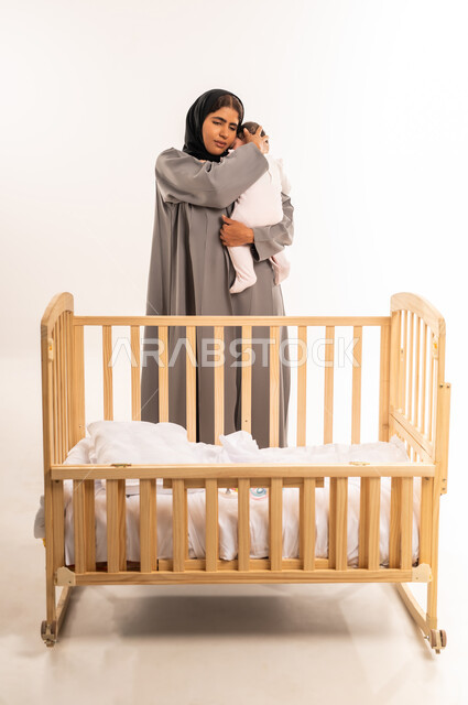 The concept of maternal tenderness, taking responsibility and caring for children, strengthening the maternal bond between mother and child, feeling safe and secure, portrait of a veiled Saudi Arabian Gulf woman wearing a gray abaya standing in front of the bed and holding her infant with expressions of love, full-length body, white background