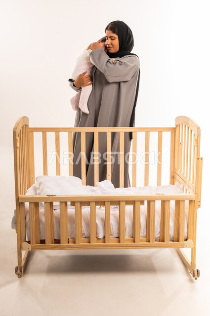 Feeling safe and secure, the concept of maternal tenderness, taking responsibility and caring for the child, strengthening the bond of motherhood between the mother and her little girl, portrait of a veiled Saudi Arabian Gulf woman wearing a gray abaya standing in front of the bed and holding her infant with expressions of love, full-length body, white background