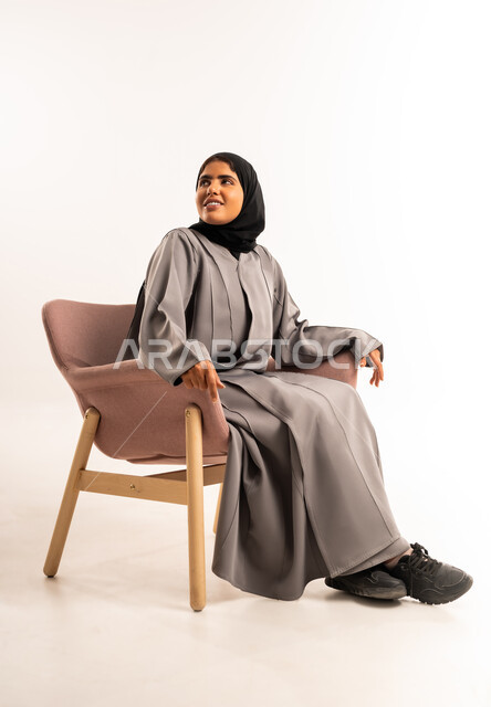 Elegance and attention, external appearance, concept of femininity and softness, portrait of a smiling veiled Saudi Arabian Gulf woman wearing a gray abaya sitting on a chair and looking at something above with gestures of happiness and pleasure, white background