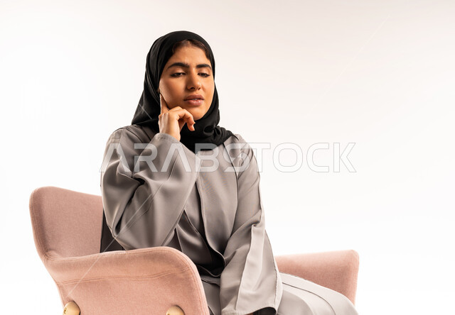 تعابير شرود الذهن والتفكير, التأمل والتخطيط للمستقبل, محاولة إيجاد حل لمشكلة ما, بورتريه لامرأة عربية خليجية سعودية محجبة ترتدي عباءة رمادية تجلس على الكرسي وتضع يدها على خدها بإيماءات التركيز والتمعن, خلفية بيضاء