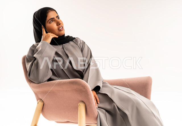 محاولة إيجاد حل لمشكلة ما, النظر للأعلى بتعابير شرود الذهن والتفكير, التأمل والتخطيط للمستقبل, بورتريه لامرأة عربية خليجية سعودية محجبة ترتدي عباءة رمادية تجلس على الكرسي وتضع يدها على خدها بإيماءات التركيز والتمعن, خلفية بيضاء
