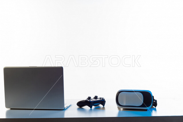 طاولة تحتوى على نظارة واقع افتراضي و عصا التحكم لألعاب الفيديو البلايستيشن ، و جهاز حاسوب محمول ، أجهزة تكنولوجية و الكترونية ، تقنيات و ألعاب ، خلفية بيضاء
