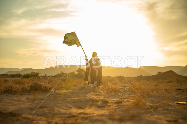 The concept of patriotism and belonging to the homeland, the day we started on February 22, celebrating the anniversary of the founding of the first Saudi state in 1727 AD, a special national occasion and holiday, wearing the central region costume, a Saudi Gulf Arab man wearing a bisht and a golden headband raising the Kingdom’s flag at sunrise in the desert