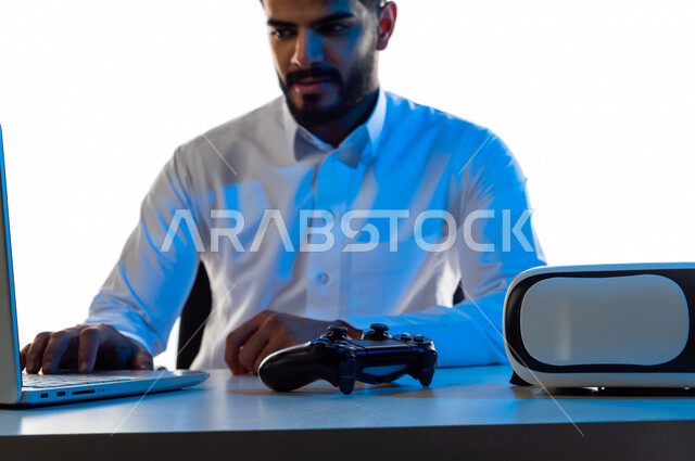 بورتريه لرجل عربي خليجي سعودي مبتسم يقوم بإستخدام الحاسوب المحمول ، يجلس على طاولة تحتوى على نظارة واقع افتراضي و عصا التحكم لألعاب الفيديو و لابتوب ، أجهزة تكنولوجية و الكترونية ، تقنيات و ألعاب ، خلفية بيضاء