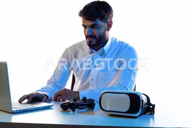 بورتريه لرجل عربي خليجي سعودي مبتسم يقوم بإستخدام الحاسوب المحمول ، يجلس على طاولة تحتوى على نظارة واقع افتراضي و عصا التحكم لألعاب الفيديو و لابتوب ، أجهزة تكنولوجية و الكترونية ، تقنيات و ألعاب ، خلفية بيضاء
