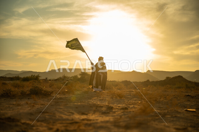 الاحتفال بمناسبة وطنية متميزة, ارتداء زي المنطقة الوسطى, الفخر والاعتزاز بالانتماء للوطن, يوم بدينا 22 فبراير, ذكرى تأسيس الدولة السعودية الأولى 1727م, رجل عربي خليجي سعودي يرتدي البشت والعقال الذهبي يرفع علم المملكة عند شروق الشمس في الصحراء