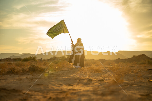 الاحتفال بذكرى تأسيس الدولة السعودية الأولى 1727م, عيد ومناسبة وطنية متميزة, ارتداء زي المنطقة الوسطى, الفخر والاعتزاز بالهوية الوطنية, يوم بدينا 22 فبراير, رجل عربي خليجي سعودي يرتدي البشت والعقال الذهبي يرفع سارية علم المملكة عند شروق الشمس في الصحراء