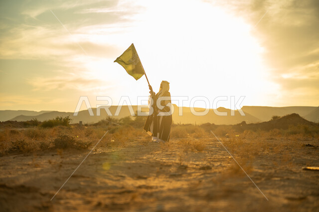 ارتداء زي المنطقة الوسطى, الفخر والاعتزاز بالهوية الوطنية, يوم بدينا 22 فبراير, الاحتفال بذكرى تأسيس الدولة السعودية الأولى 1727م, مناسبة وعيد وطني متميز, رجل عربي خليجي سعودي يرتدي البشت والعقال الذهبي يرفع سارية علم المملكة عند شروق الشمس في الصحراء
