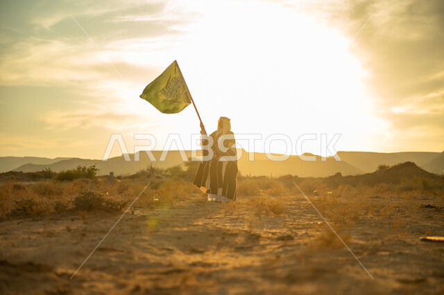 الفخر والاعتزاز بالهوية الوطنية, يوم بدينا 22 فبراير, الاحتفال بذكرى تأسيس الدولة السعودية الأولى 1727م, مناسبة وعيد وطني متميز, ارتداء زي المنطقة الوسطى, رجل عربي خليجي سعودي يرتدي البشت والعقال الذهبي يرفع سارية علم المملكة عند شروق الشمس في الصحراء