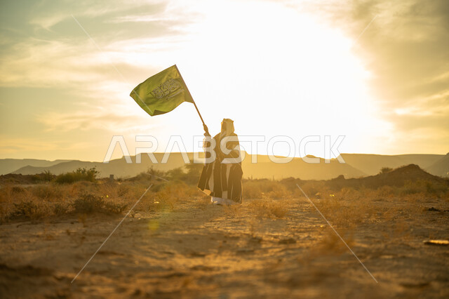 الاعتزاز والفخر بالهوية الوطنية, يوم بدينا 22 فبراير, الاحتفال بذكرى تأسيس الدولة السعودية الأولى 1727م, مناسبة وعيد وطني متميز, ارتداء زي المنطقة الوسطى, رجل عربي خليجي سعودي يرتدي البشت والعقال الذهبي يرفع سارية علم المملكة عند شروق الشمس في الصحراء